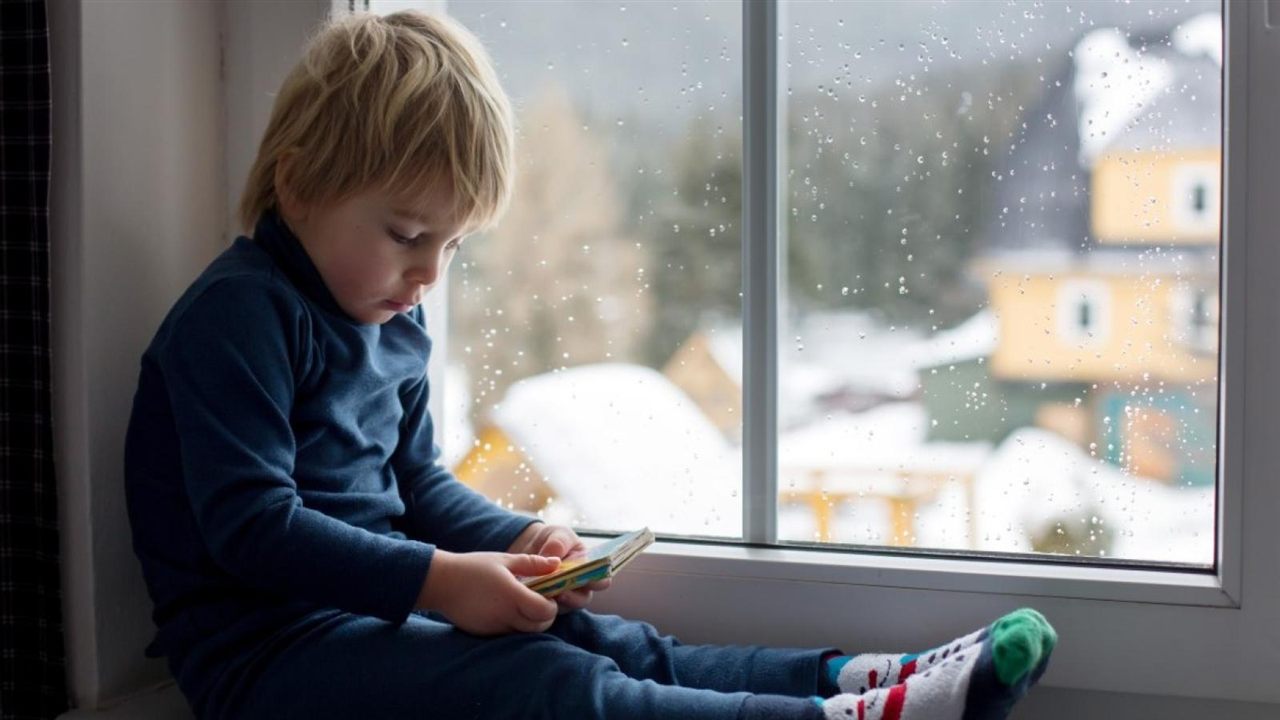 A young child sitting on a windowsill on a snowy day, looking at a book