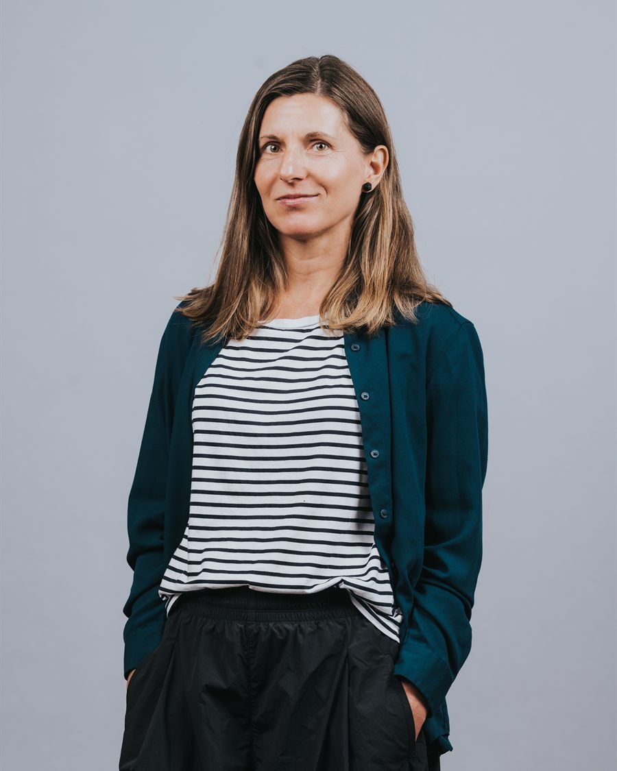 Portrait of Nataliya Deleva, standing with her hands in her pockets, wearing a striped top and teal cardigan